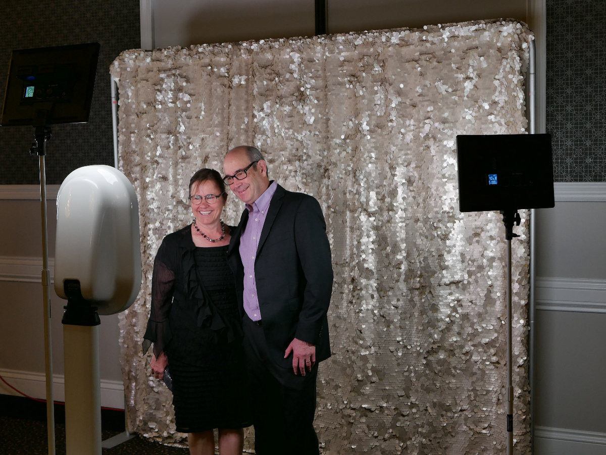 A man and woman pose together in front of a digital photobooth screen. Behind them is a gold sequin backdrop,